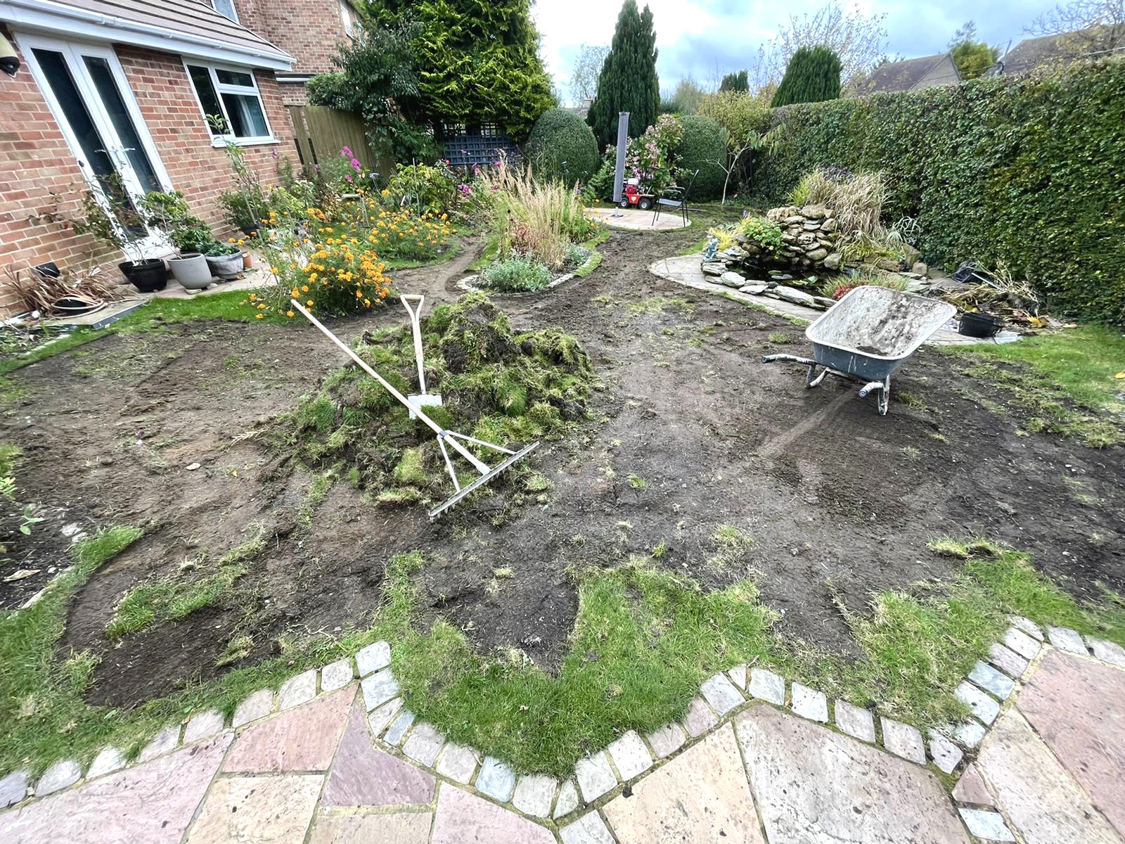 Rear garden stripped back to bare soil before turfing, with a wheelbarrow and the cobble-edged patio